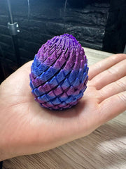 Acusinkt Drakenei in iriserend paars-blauw-roze, gesloten ei op een volwassen hand ter vergelijking van de grootte.